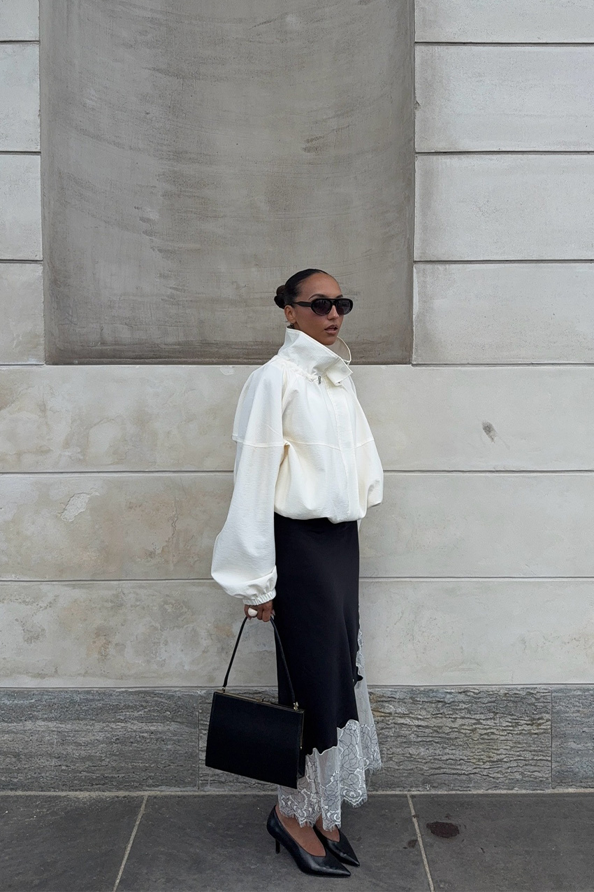 Trendy basics at Nordstrom are shown in a street style photo of a woman standing on a sidewalk wearing black sunglasses, a white funnel neck jacket, a black slip skirt with contrasting white lace trim, black pointed pumps, a black frame handbag, and a white resin ring