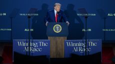 US President Donald Trump speaking at a podium during the "Winning the AI Race" summit, flanked by the slogans "Winning The AI Race" and "ALL-IN" on boards.