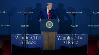 US President Donald Trump speaking at a podium during the "Winning the AI Race" summit, flanked by the slogans "Winning The AI Race" and "ALL-IN" on boards.