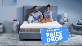 A man and woman sit on the Tempur-Pedic Tempur-Cloud mattress on an upholstered bed frame in a bedroom. A Tom's Guide Price Drop deals badge is in the bottom right corner