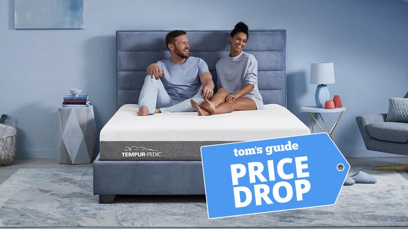 A man and woman sit on the Tempur-Pedic Tempur-Cloud mattress on an upholstered bed frame in a bedroom. A Tom&#039;s Guide Price Drop deals badge is in the bottom right corner