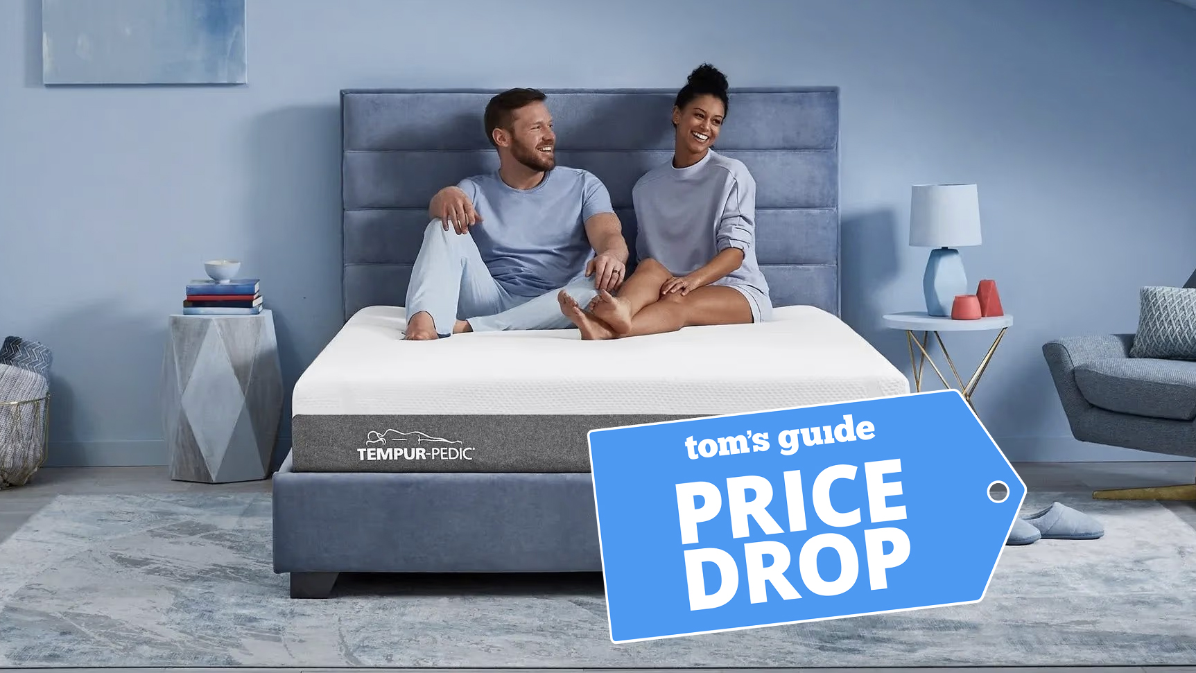 A man and woman sit on the Tempur-Pedic Tempur-Cloud mattress on an upholstered bed frame in a bedroom. A Tom&#039;s Guide Price Drop deals badge is in the bottom right corner