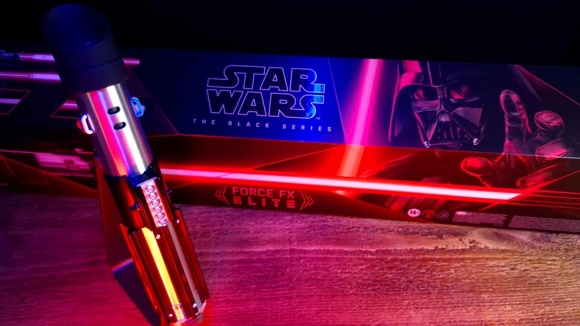 The Darth Vader Force FX Elite Lightsaber on a stand beside its box, all on a wooden table in a darkened space