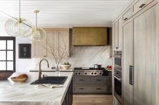 A modern kitchen with taupe shaker style cabinetry, a metallic gold toned oven vent, and a large marble island