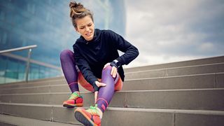 Woman with a pained expression and wearing running clothes sits on steps holding her leg