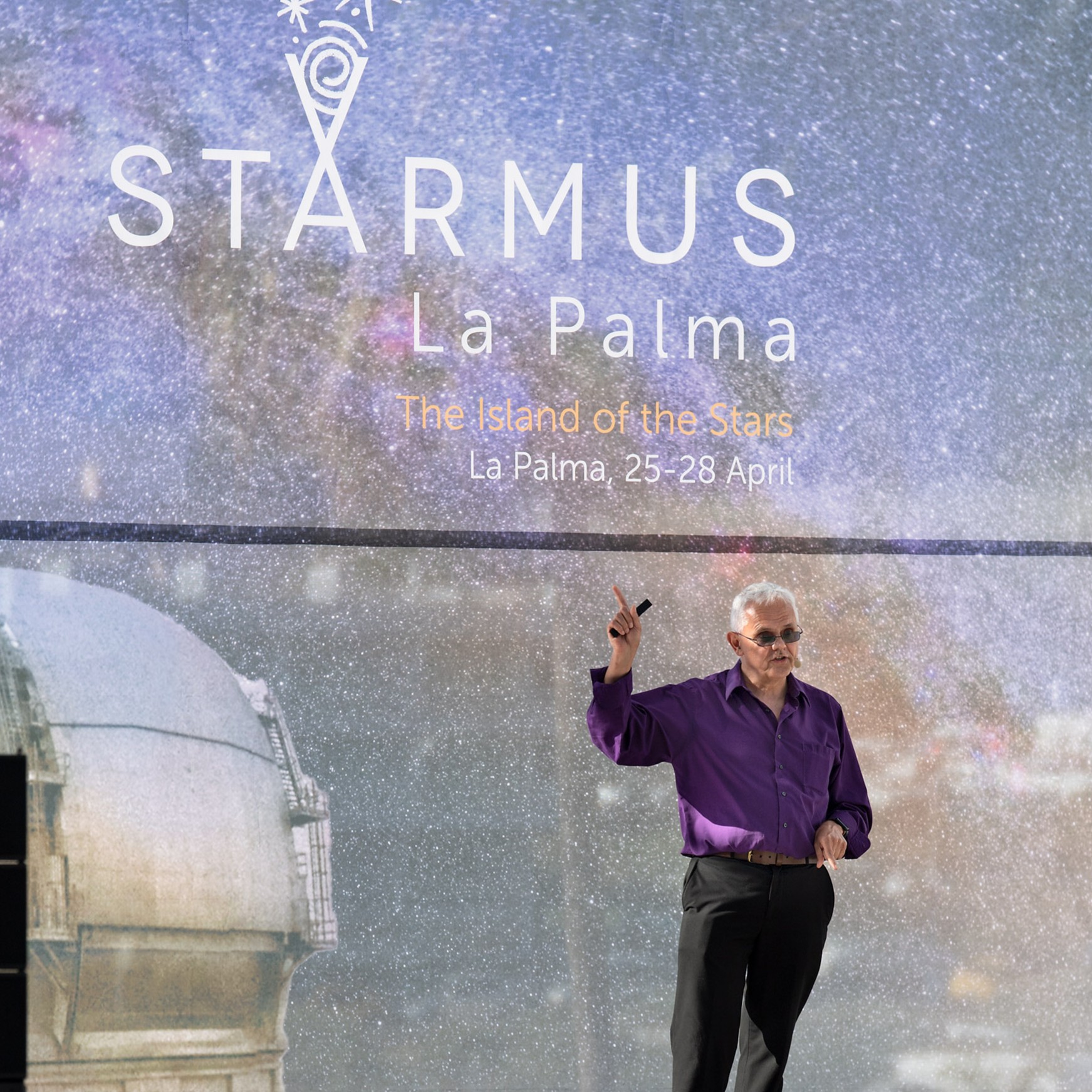 a man stands on a stage with STARMUS La Palma written on a fabric backdrop behind.