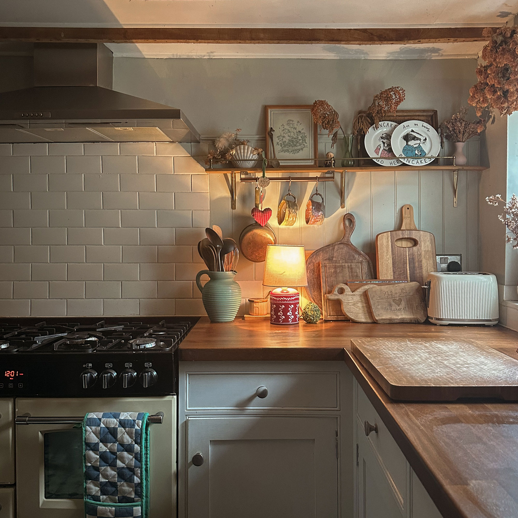 Corner of kitchen with wooden countertops, small lamp, several wooden chopping boards as well as mugs hanging from brass rail