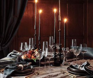 A dark dinner party scene, with tall black tapered candles, wine glasses and plates.