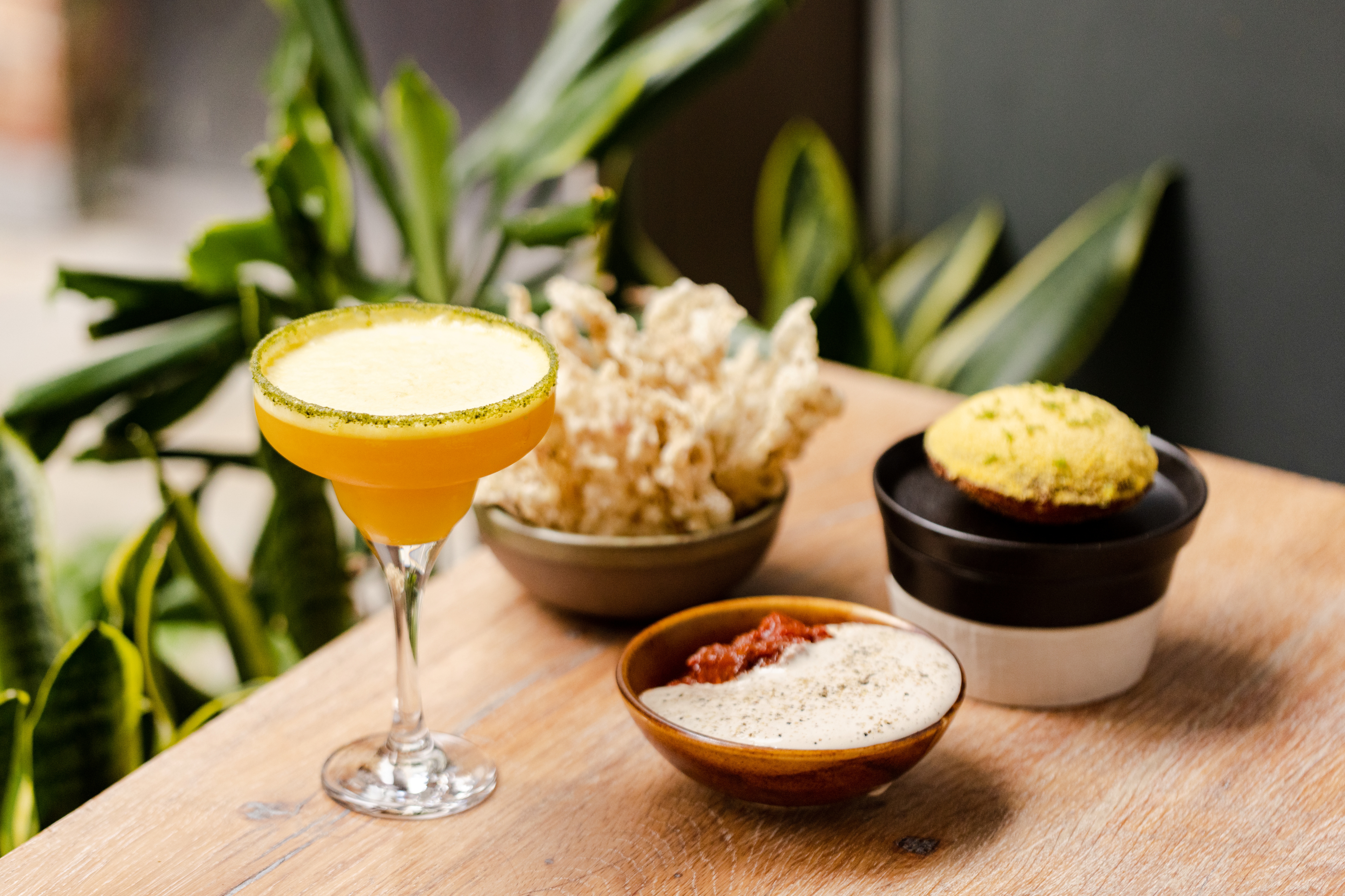 A pale wood table covered in multiple sharing platters filled with colorful food and drinks is placed next to a series of indoor plants.