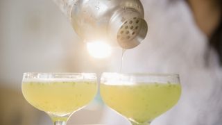 two filled coupe glasses in front of a woman pouring cocktails into one of them from a shaker