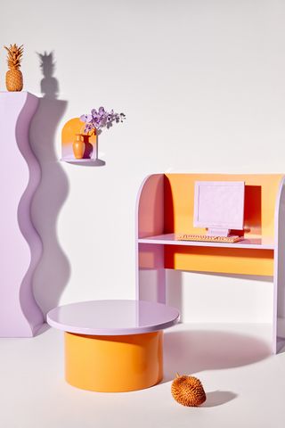 A study furniture setup has lilla and orange-shaped furniture including a wavy wardrobe, a desk, and a coffee table.