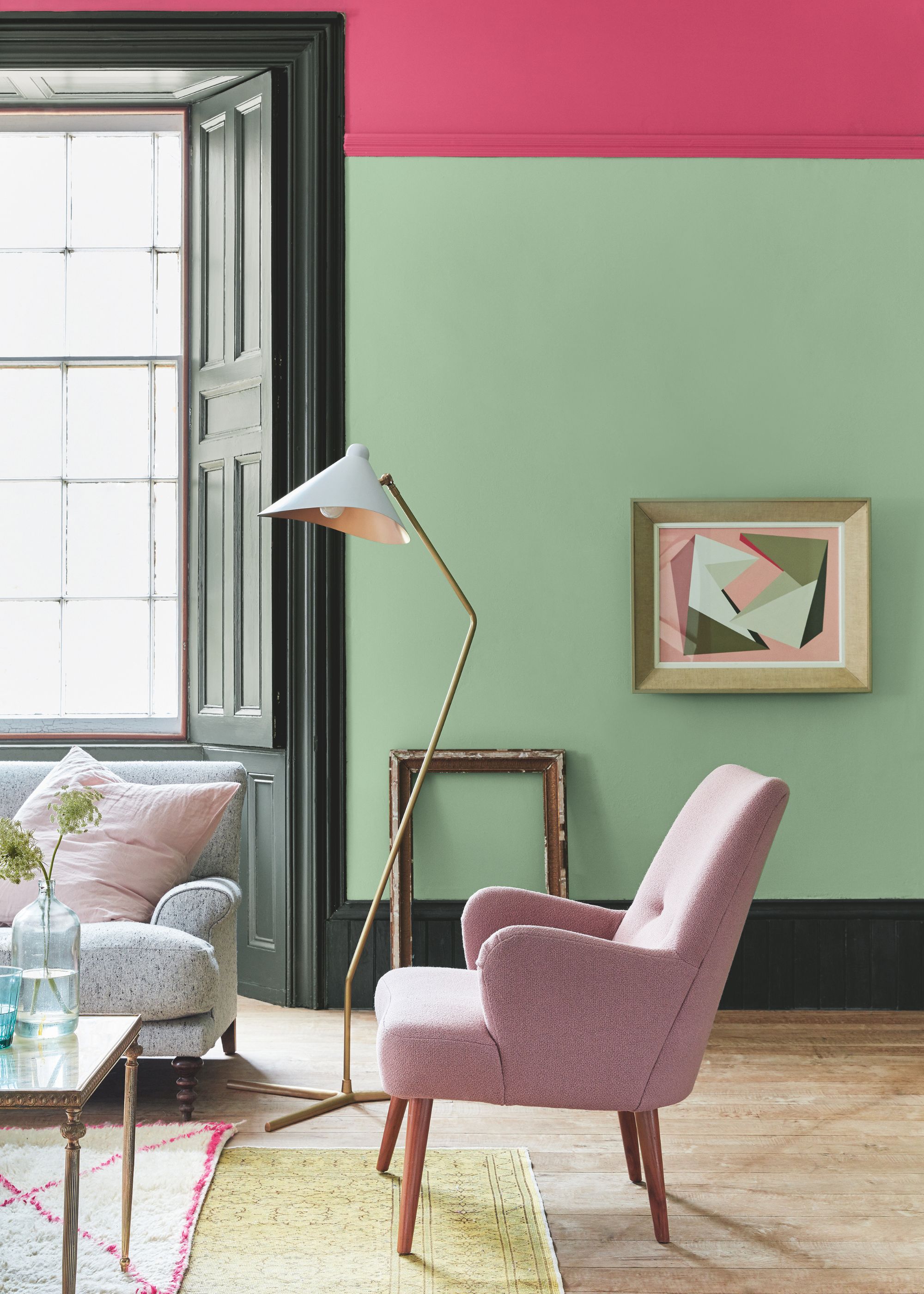 A living room with light green walls and dark pink upper portion of wall, light pink armchair, and large window with dark green woodwork.