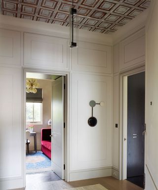 Hallway with white painted walls and large rectangular panels, ceiling painted in white and brown to create a painted paneling effect, wooden flooring and natural, textured rug, industrial style hanging pendant and metallic circular decorative feature hanging on wall