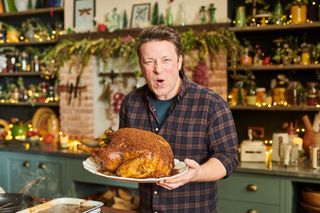 Jamie Oliver holds up a freshly-cooked Christmas turkey, browned to perfection and covered in a flavoursome rub. He is looking into the camera with a delighted expression