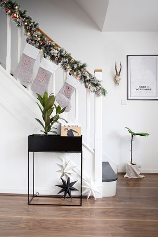 White staircase decorated with a green frosted evergreen and pine cone Christmas garland, grey stockings and lights. Black console table with plants and paper stars