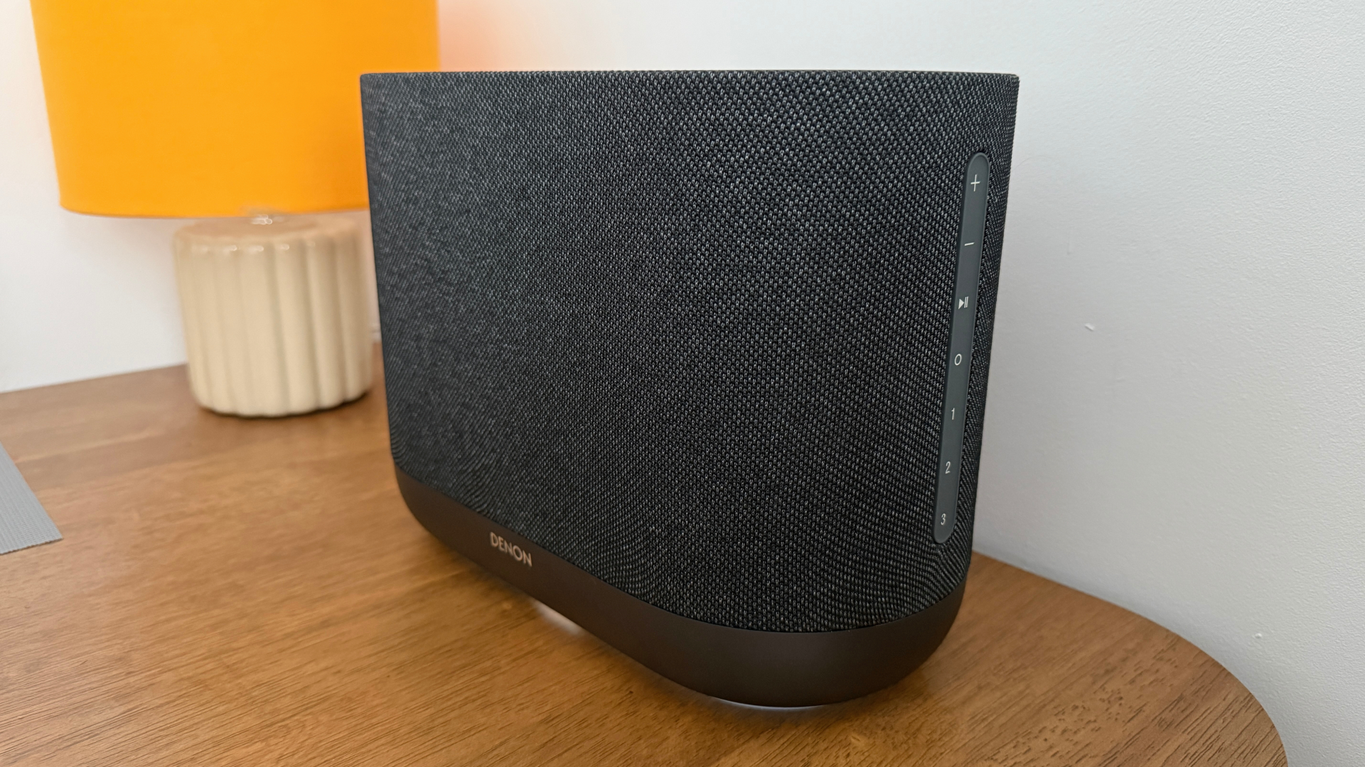 A side-on view of a Denon Home 400 wireless speaker. It's positioned on a wooden table with a lamp in the background.