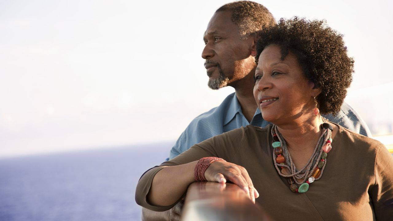 An older, relaxed-looking couple stand together at the railing of a cruise ship. 