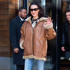 Bella Hadid wears flared jeans with a brown leather jacket and sunglasses.