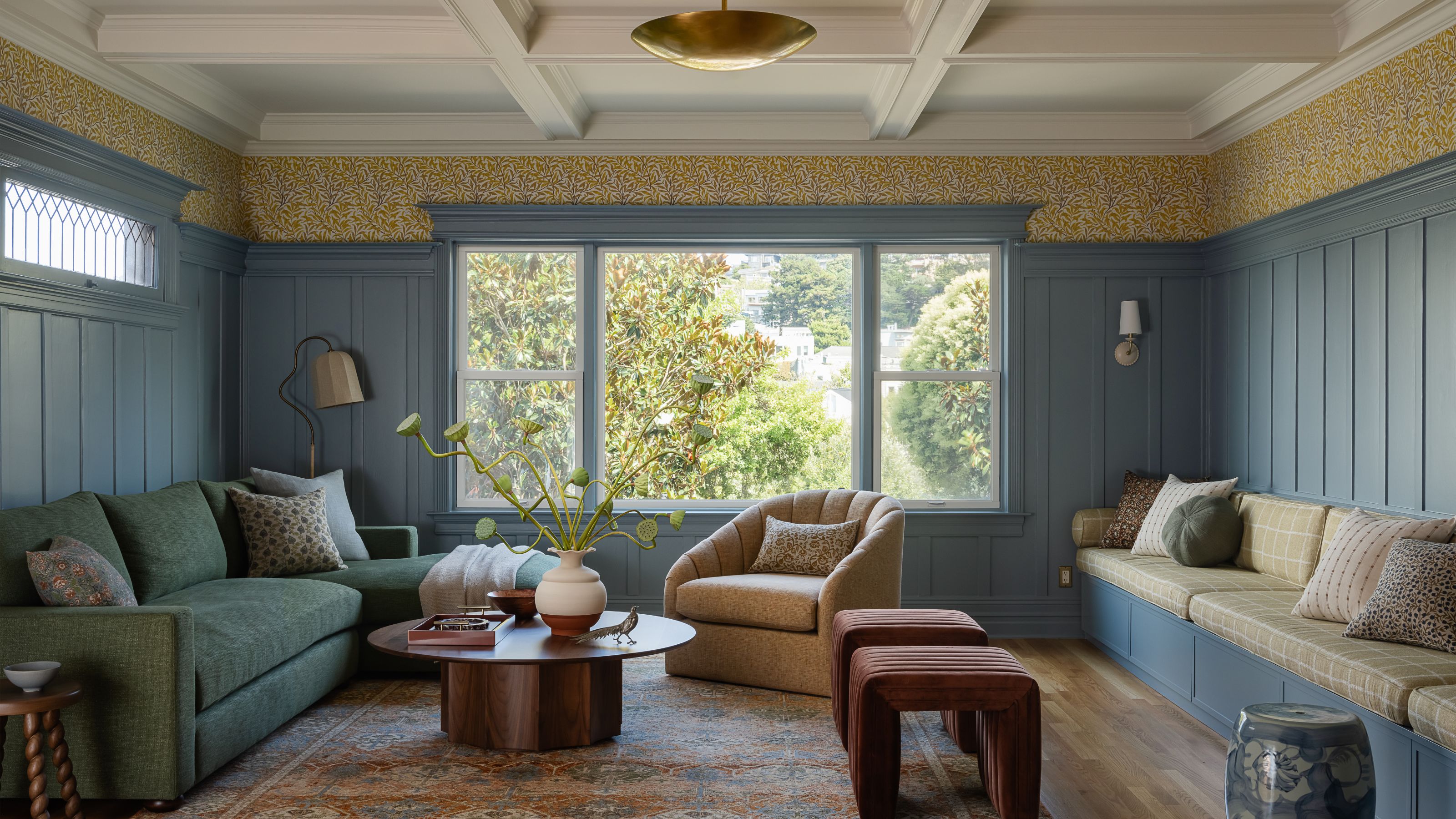 A living room with blue paneled walls, yellow floral wallpaper, a green sofa, and a built-in window seat, centered around a round wood coffee table.