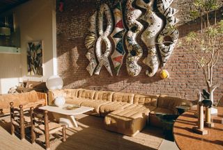 A sun-kissed, open space area features organic brown, leathery couches and wooden chairs and tables while large-scale, colorful art pieces hang on the brick walls of the room.