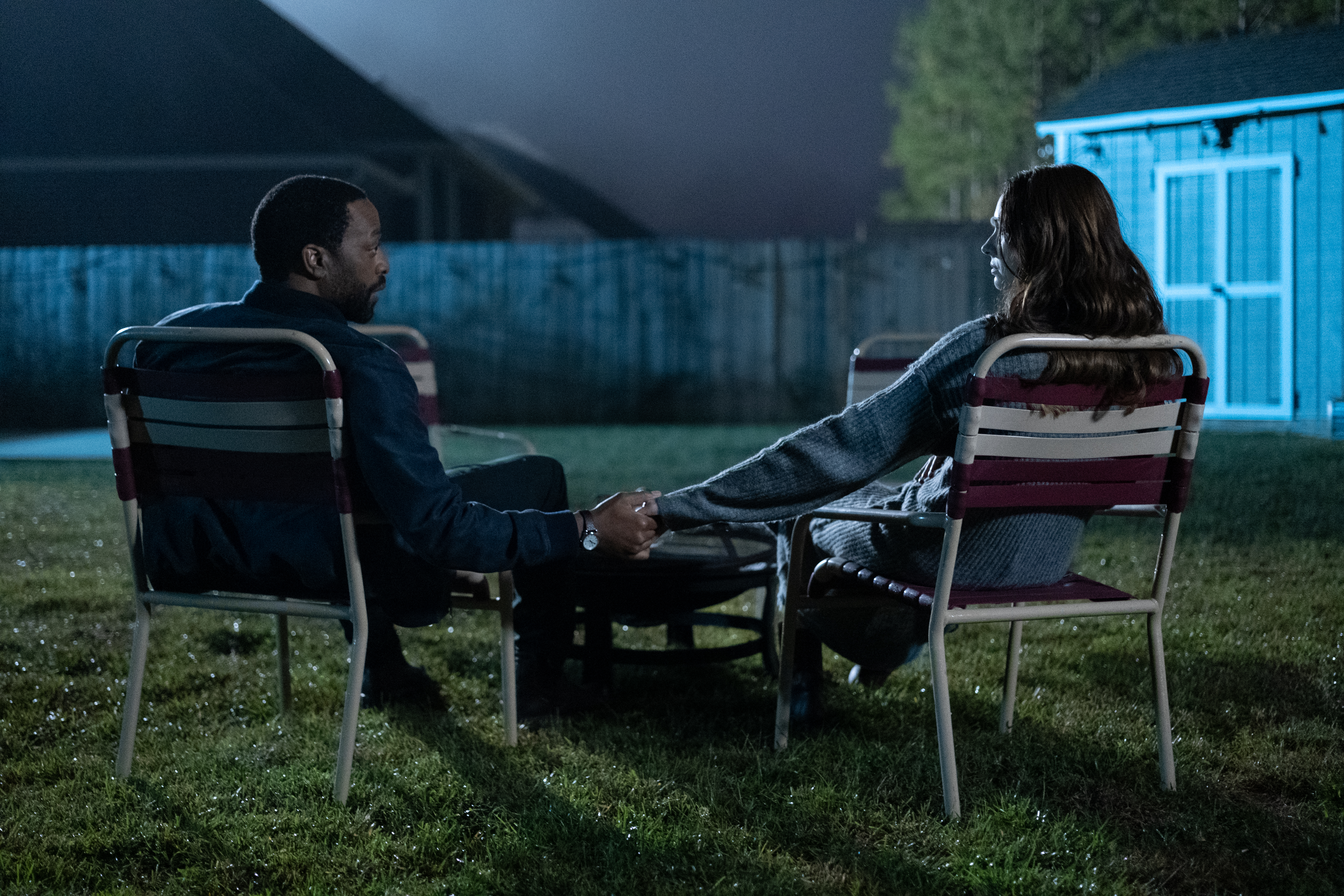 A Black man (actor Chiwetel Ejiofor) and a white woman (actor Karen Gillan) sit in lawn chairs, holding hands in front of a circular unlit fire pit. They sit in a suburban backyard at night, with a blue glow illuminating a fence and a wooden shed. A still from 'The Life of Chuck.'