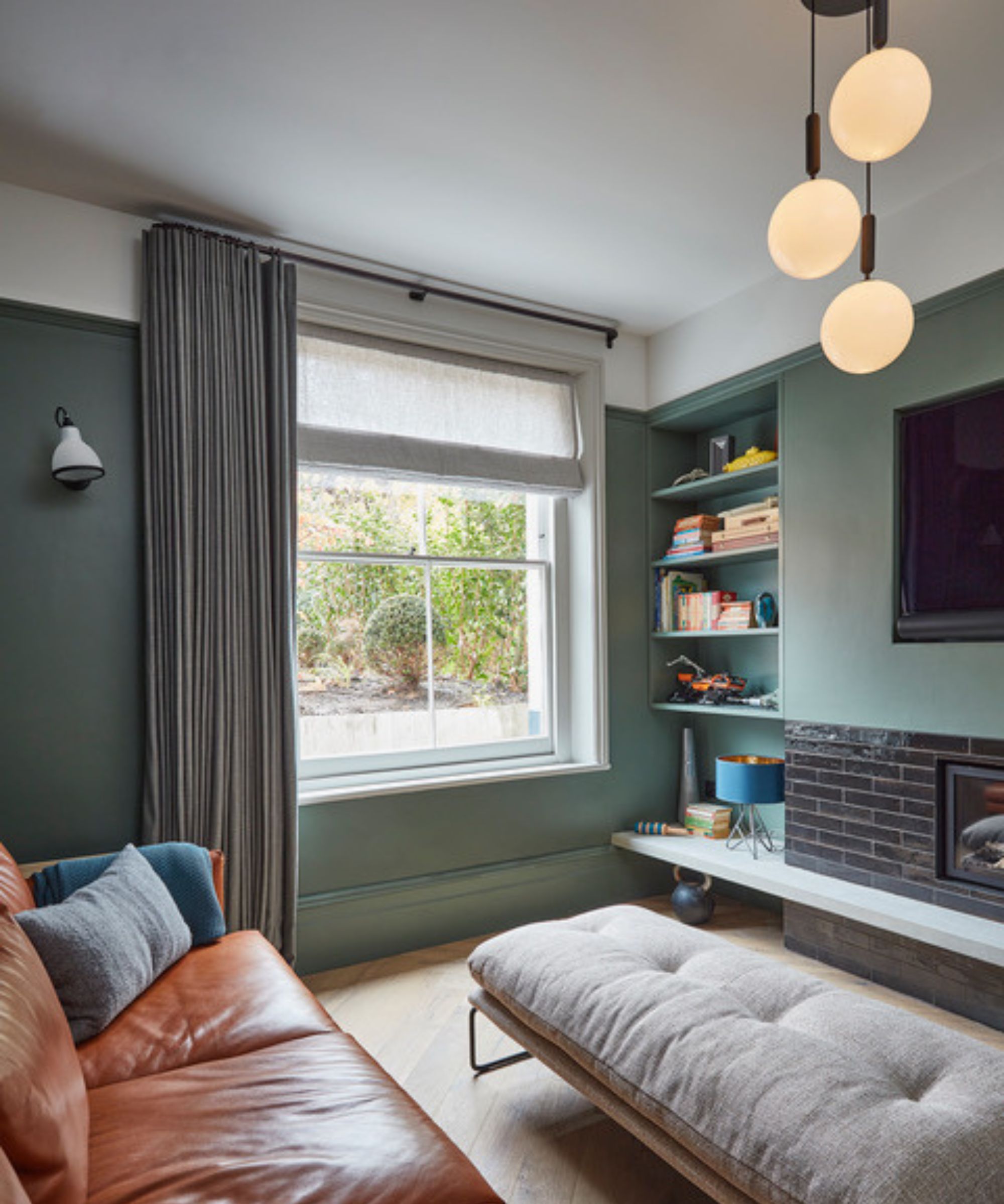 A compact living room with a contemporary feel. The space features green-painted walls with built-in shelving, a sleek grey curtain framing a sash window, and a brown leather sofa paired with a cushioned bench-style seat. A cluster pendant light with three round globes hangs from the ceiling.