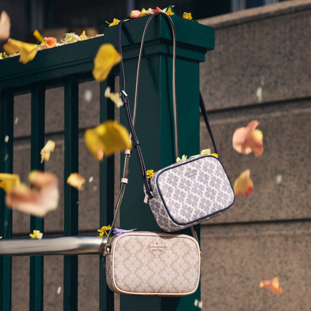 Two pattered Kate Spade handbags hanging on a green fence with autumn leaves falling in the foreground