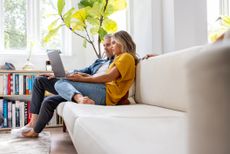 A couple in their late 50s or early 60s relaxes on their couch at home. They are looking at a laptop together and seem to be concentrating.