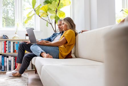 A couple in their late 50s or early 60s relaxes on their couch at home. They are looking at a laptop together and seem to be concentrating.