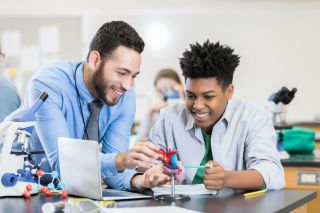 Teenage boy in High School science lab studies model of the human heart, while male teacher manipulates the model to answer his questions.