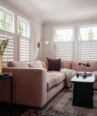 white living room with shutters, pale pink sectional, pink vintage rug, black wood coffee table, burgundy couch pillows, wall light,