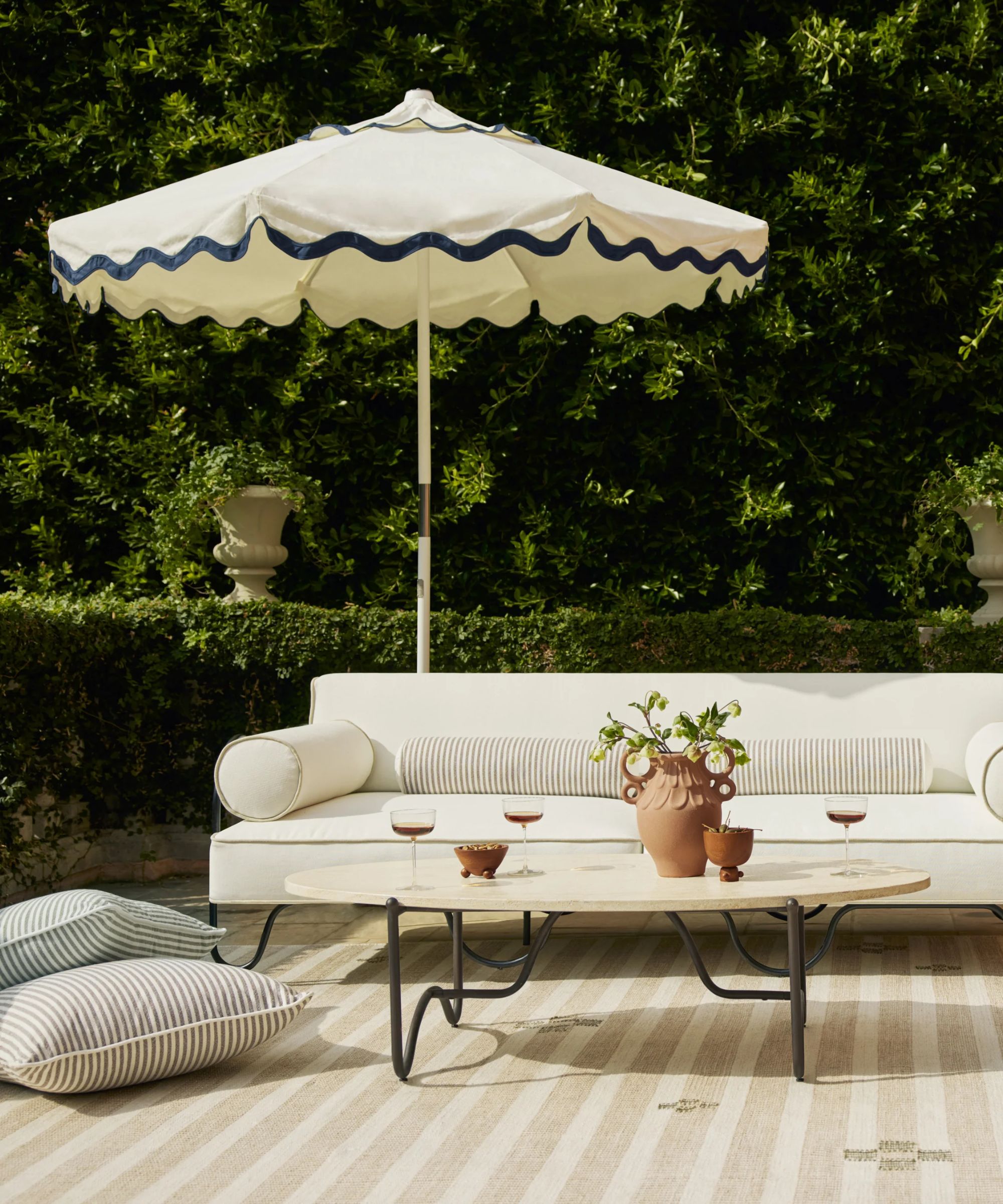 patio area surrounded by green hedges with black metal furniture, white cushions, striped throw pillows, and a white and black scalloped umbrella