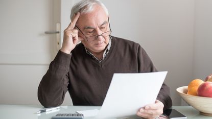 An older man sits at a kitchen counter with a calculator in front of him and looks at paperwork with a thoughtful expression.