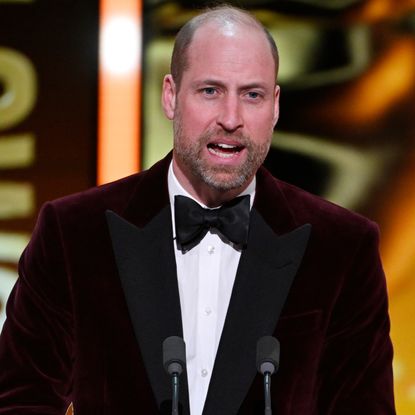 Prince William wearing a burgundy jacket and tie speaking in a mic