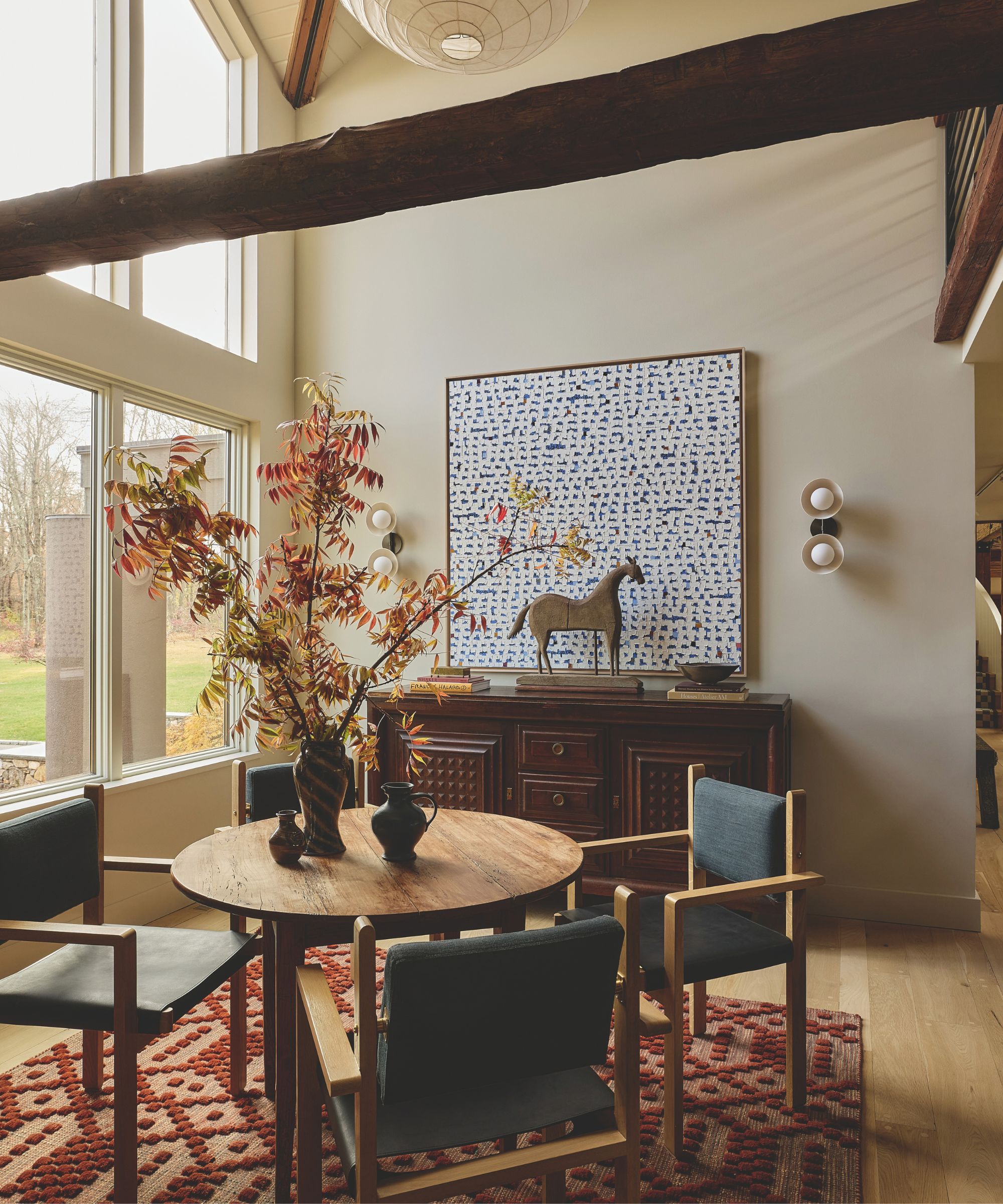 Round wooden dining table with gray armchairs under exposed ceiling beams, abstract wall art, and tall autumn branches in a vase.