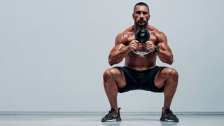 Man in a squat position holding a kettlebell with both hands against grey backdrop