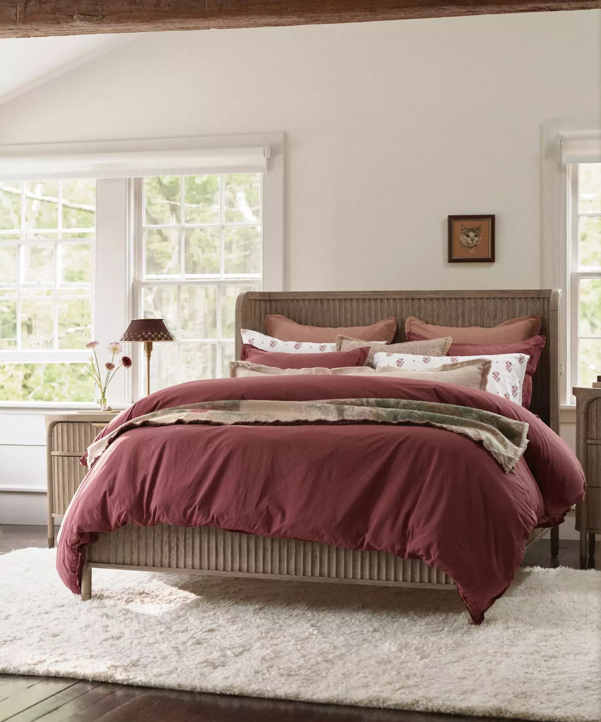 White bedroom with bed, wooden headboard, berry-red bedding and throw pillows, cream furry rug, artwork on wall