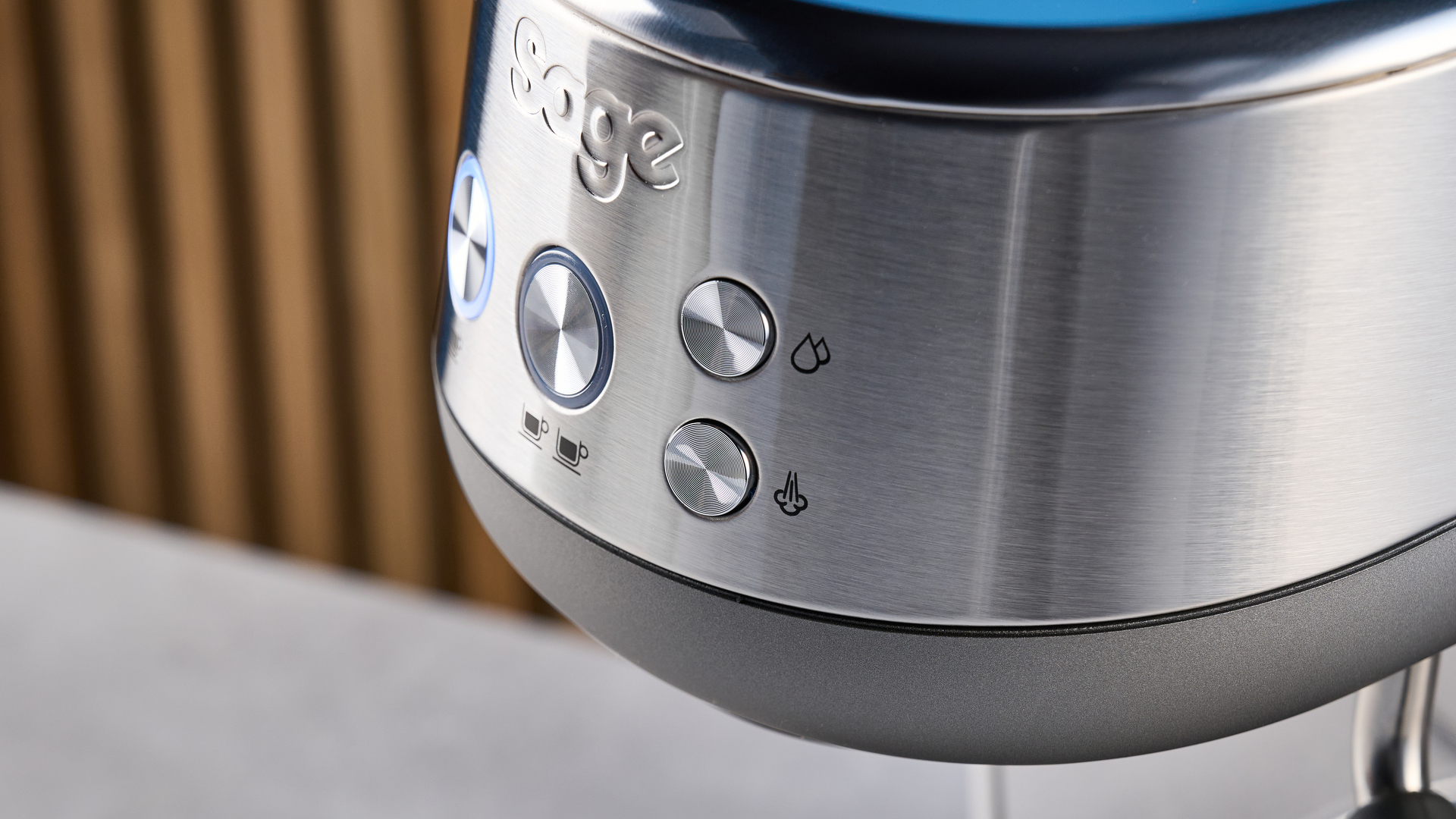 the breville bambino / sage bambino in silver photographed against a blue background with a wooden feature wall, showing the steam wand, group head, water tank, button controls, and espresso