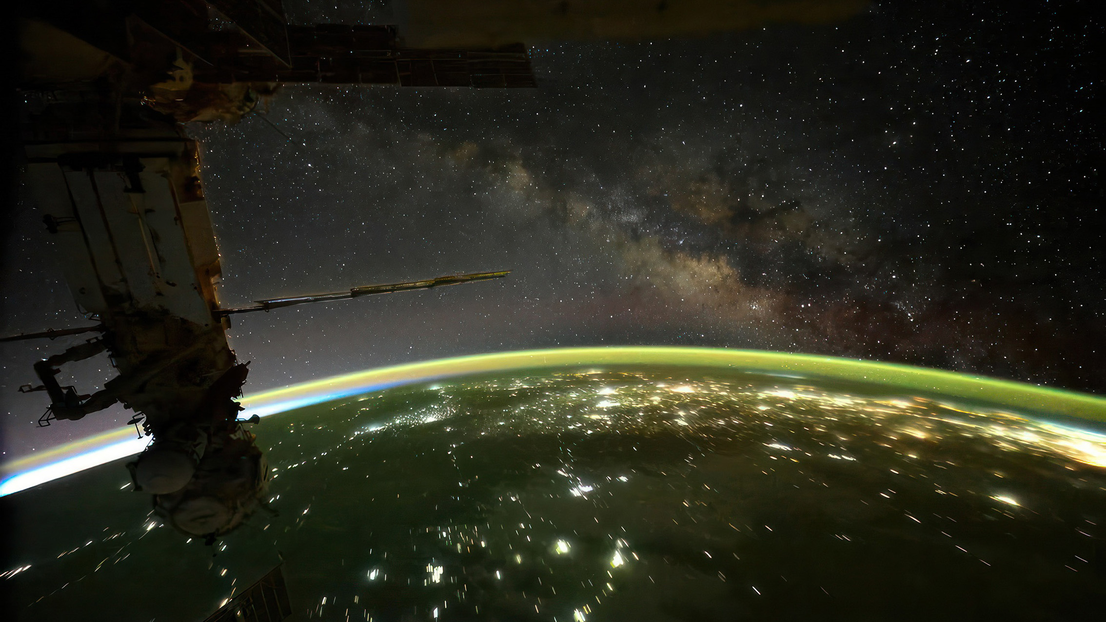 An aerial view from the ISS shows Earth illuminated at night, with a vibrant atmosphere and the Milky Way stretching across the starry sky