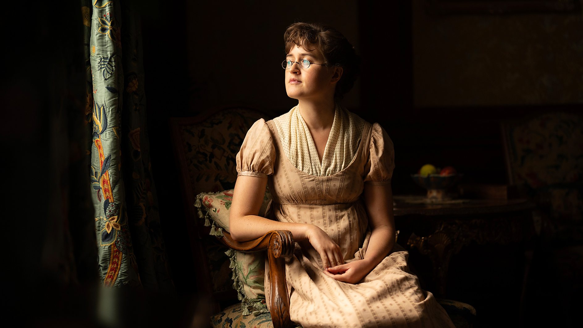 A Regency-era woman (Ella Bruccoleri as Mary Bennet) sits in a wooden chair next to a curtained window, with sunlight illuminating a darkened room, in BritBox's 'The Other Bennet Sister.'