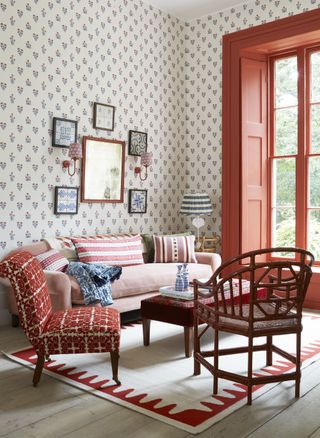 apartment idea with small print wallpaper, orange painted woodwork around window, pink couch, red chairs and coffee table, cream and red rug, whitewashed floor boards, artwork, wall lights, table lamp