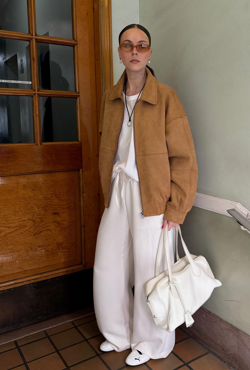 The best ballet sneakers are shown in a photo of a woman standing in a doorway wearing a tan suede jacket over a white t-shirt with white sweatpants and white ballet sneakers and a white bowler bag
