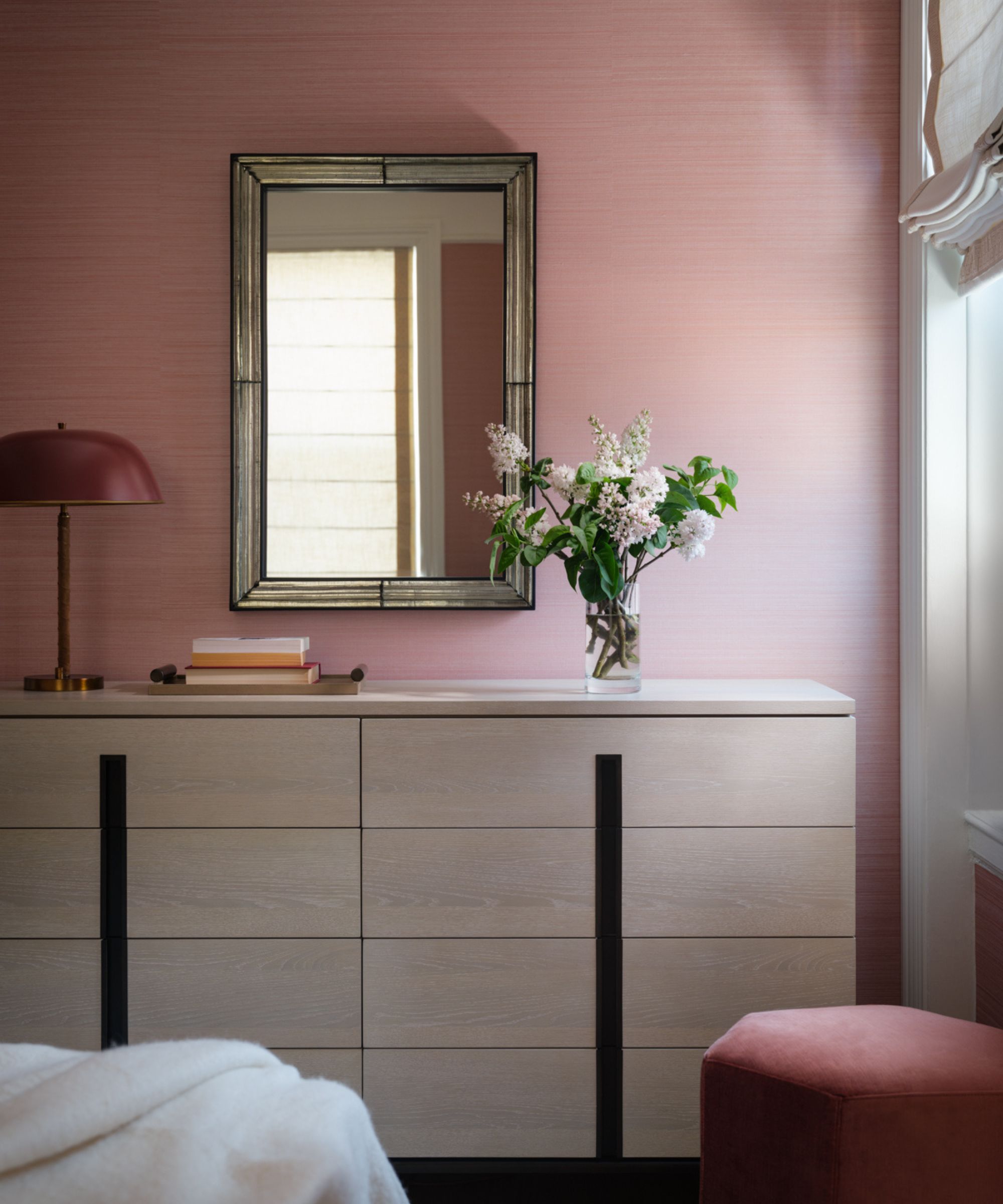 The corner of a bedroom with earthy pink textured wallpaper, a white wooden modern dresser with a vase of flowers, a red lamp, and a mirror above it. A dusky pink footstool and a window with Roman blinds.