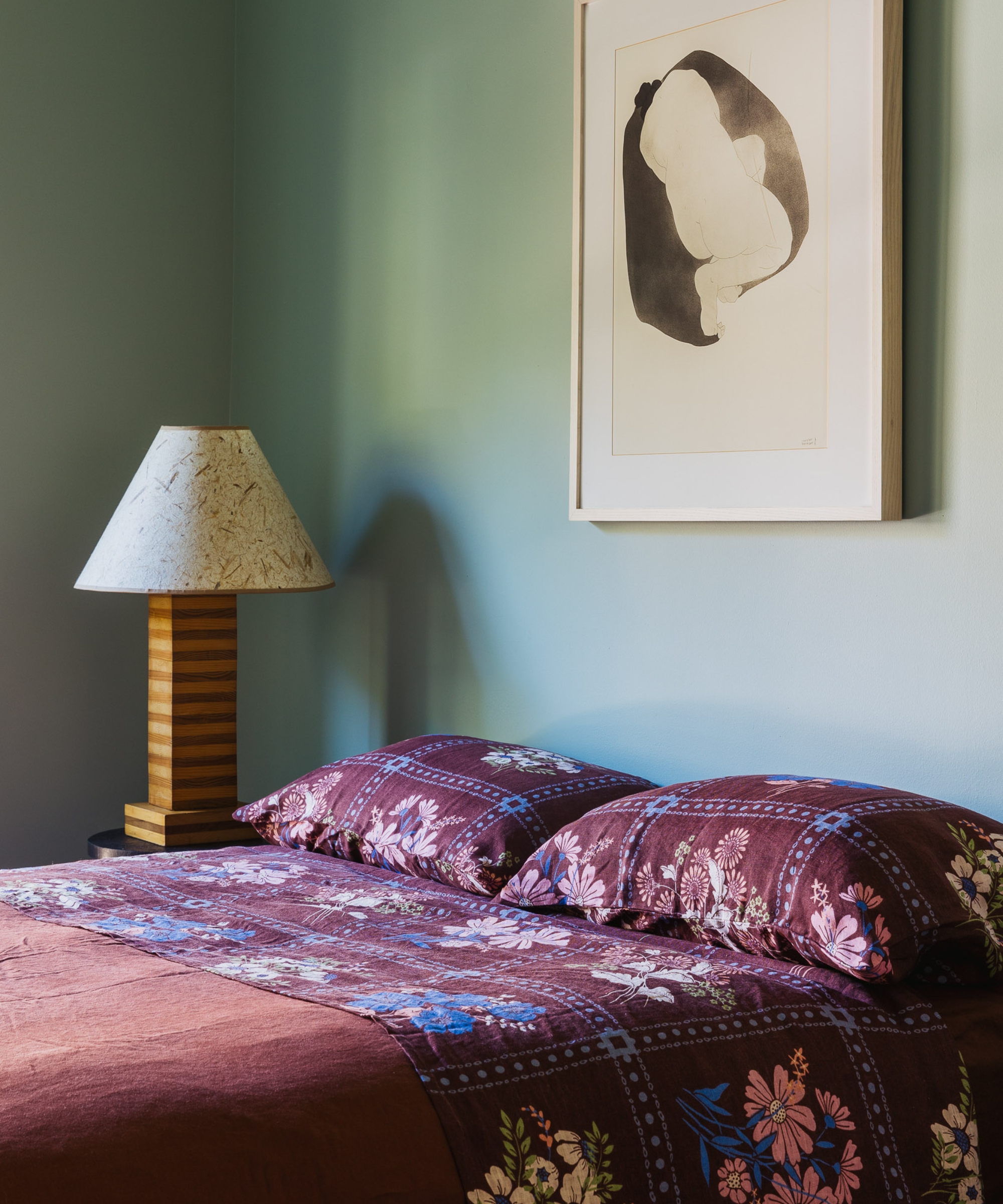 Bedroom with soft blue-green walls, burgundy floral bedding, a striped wood lamp and framed monochrome artwork.