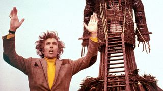 edward woodward raising his arms up while standing in front of a wood statue in wicker man