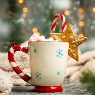 A red and white Christmas mug with marshmallows and a candy cane in a Christmas scene.