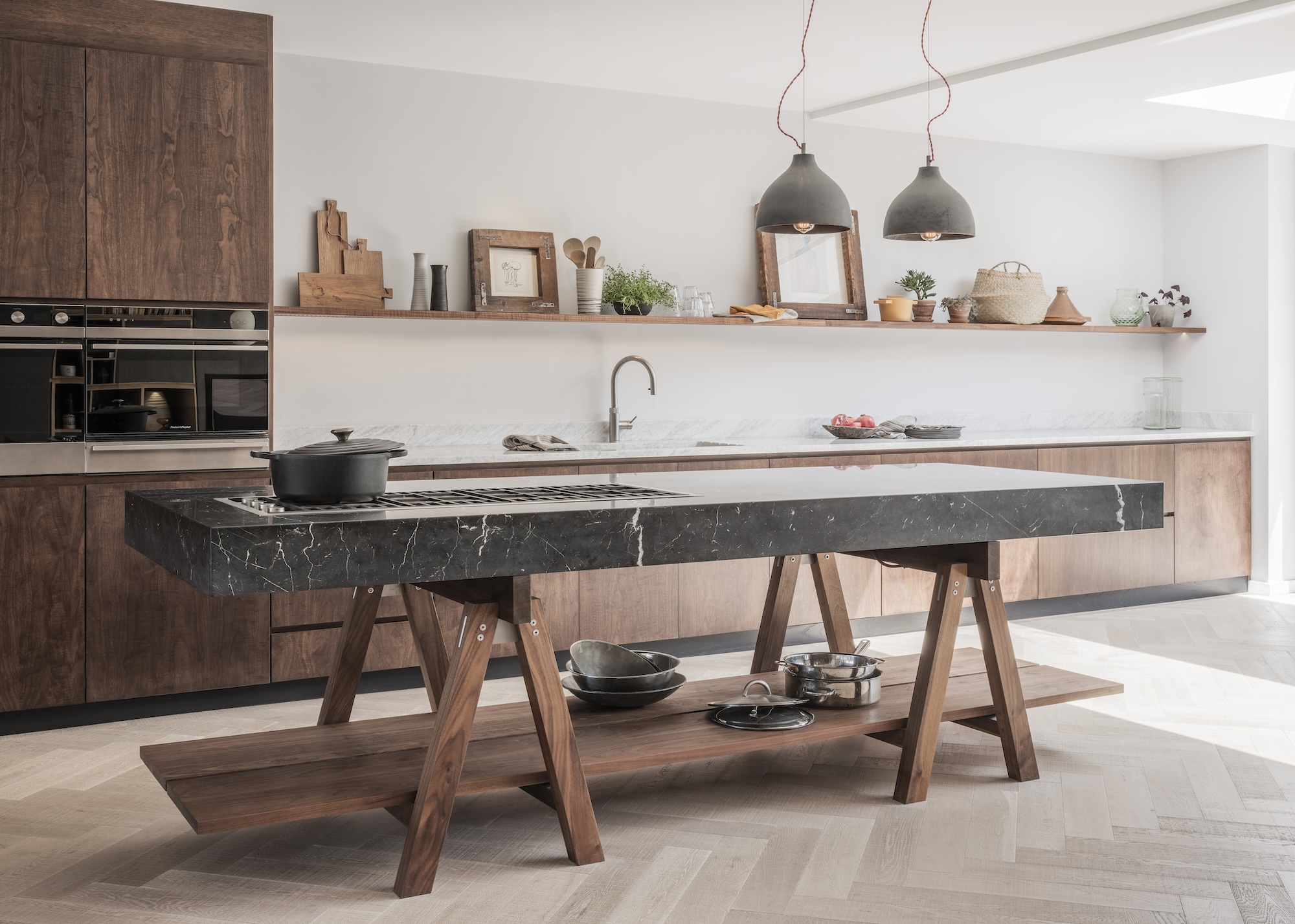 A large table/portable island in the middle of a kitchen with a stove on top of it as well as open shelving at the bottom. There is also a view of the rest of the kitchen which has open shelving and wooden textures and accessories.