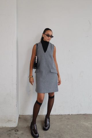 A woman standing in a garage wearing black sunglasses, a black sleeveless turtleneck under a gray v-neck dress, black knee-high stockings, black loafers, a black handbag, and a silver bangle.