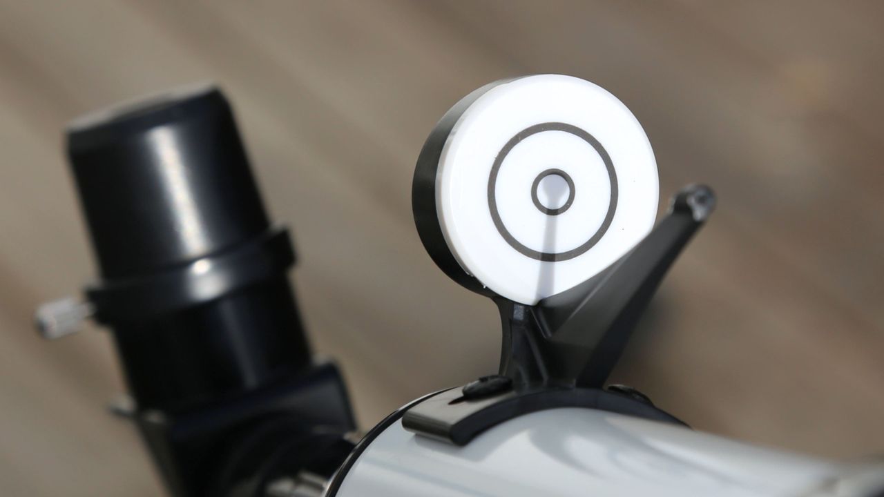 A white circle with interior black circles making a target is attached to a white curved base of a telescope. A black telescope eyepiece is seen in the background.