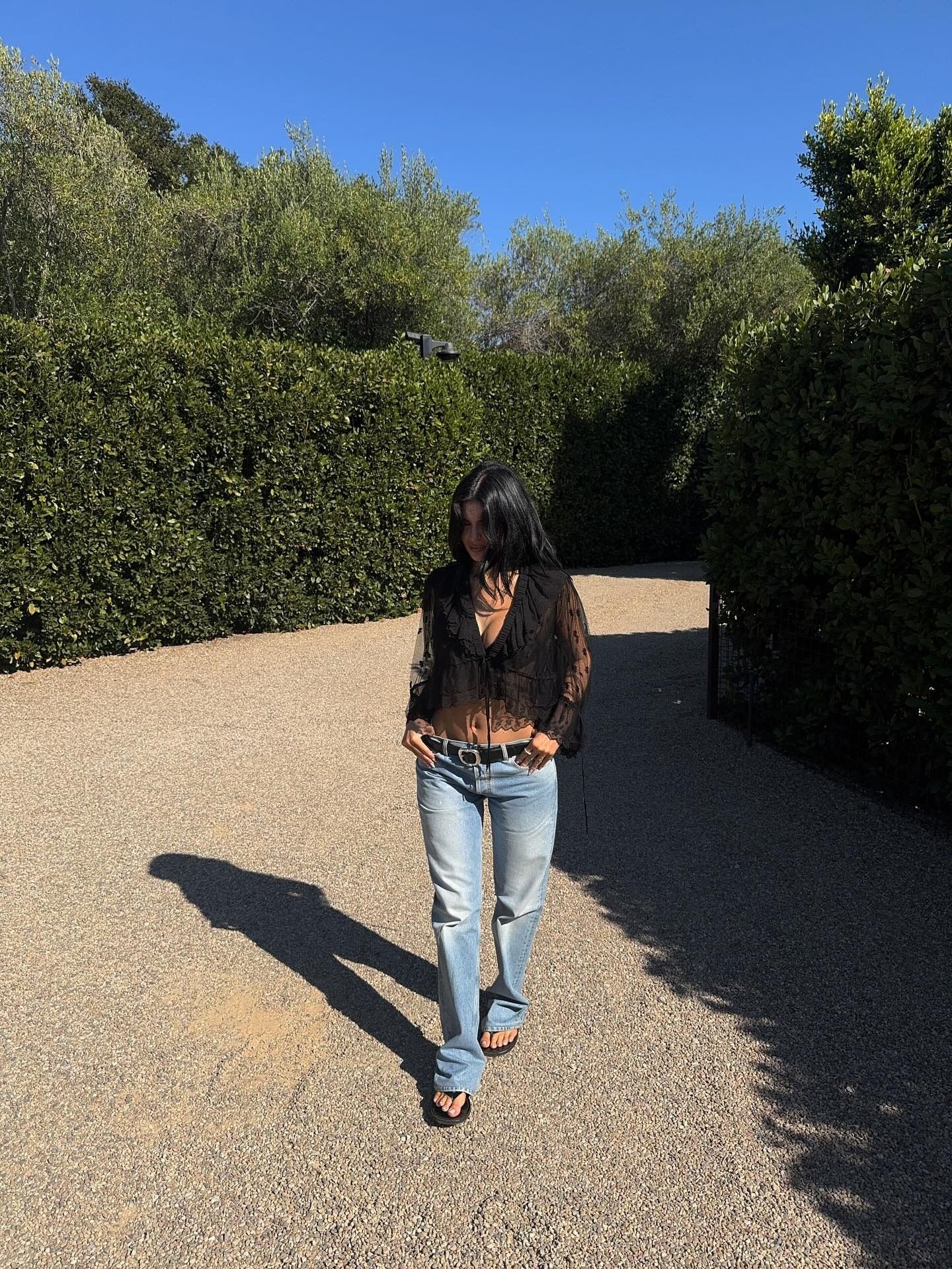 Kylie wears a black lace blouse, black belt, low-rise jeans, and black flip-flops.